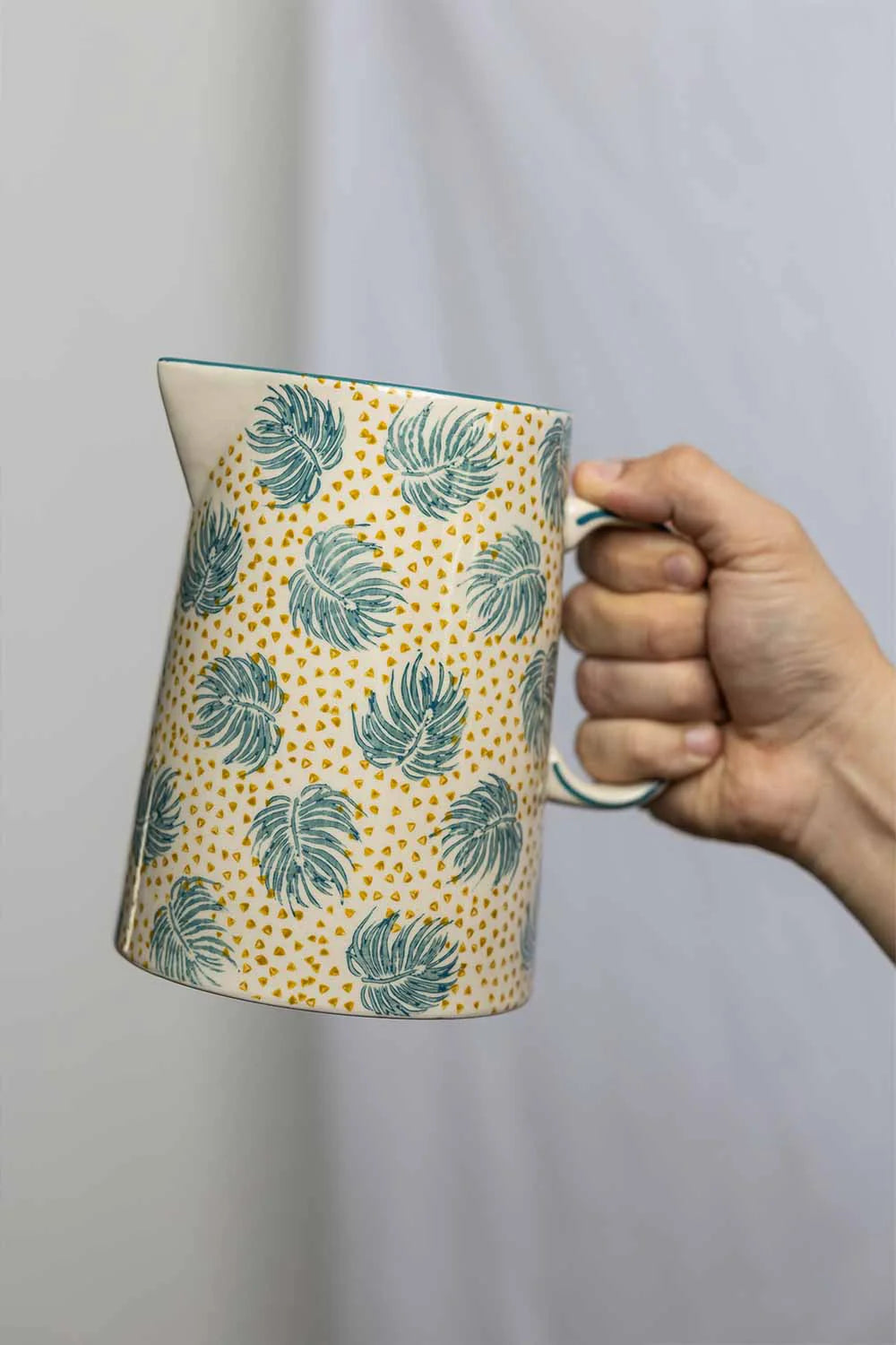 Hand holding a ceramic pitcher with leaf pattern against a plain background
