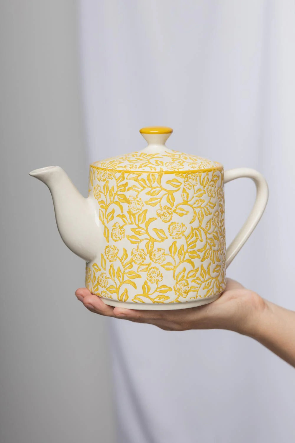 White teapot with yellow floral pattern held by a hand against a gray background