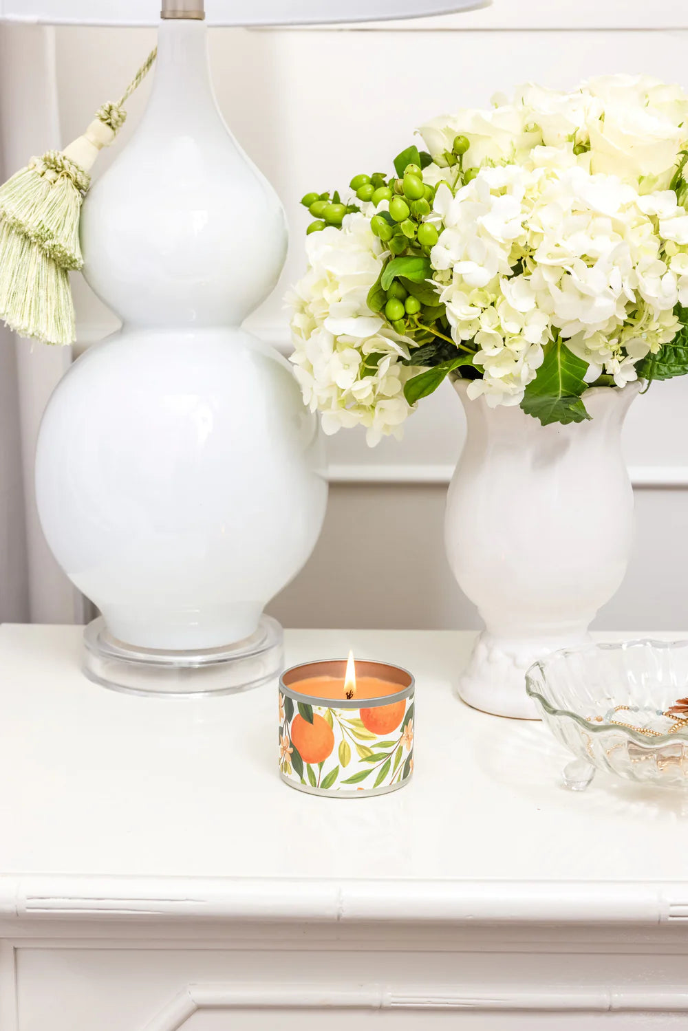 lit Orange & Honey Tin Candle set on a side table with white vases around it.
