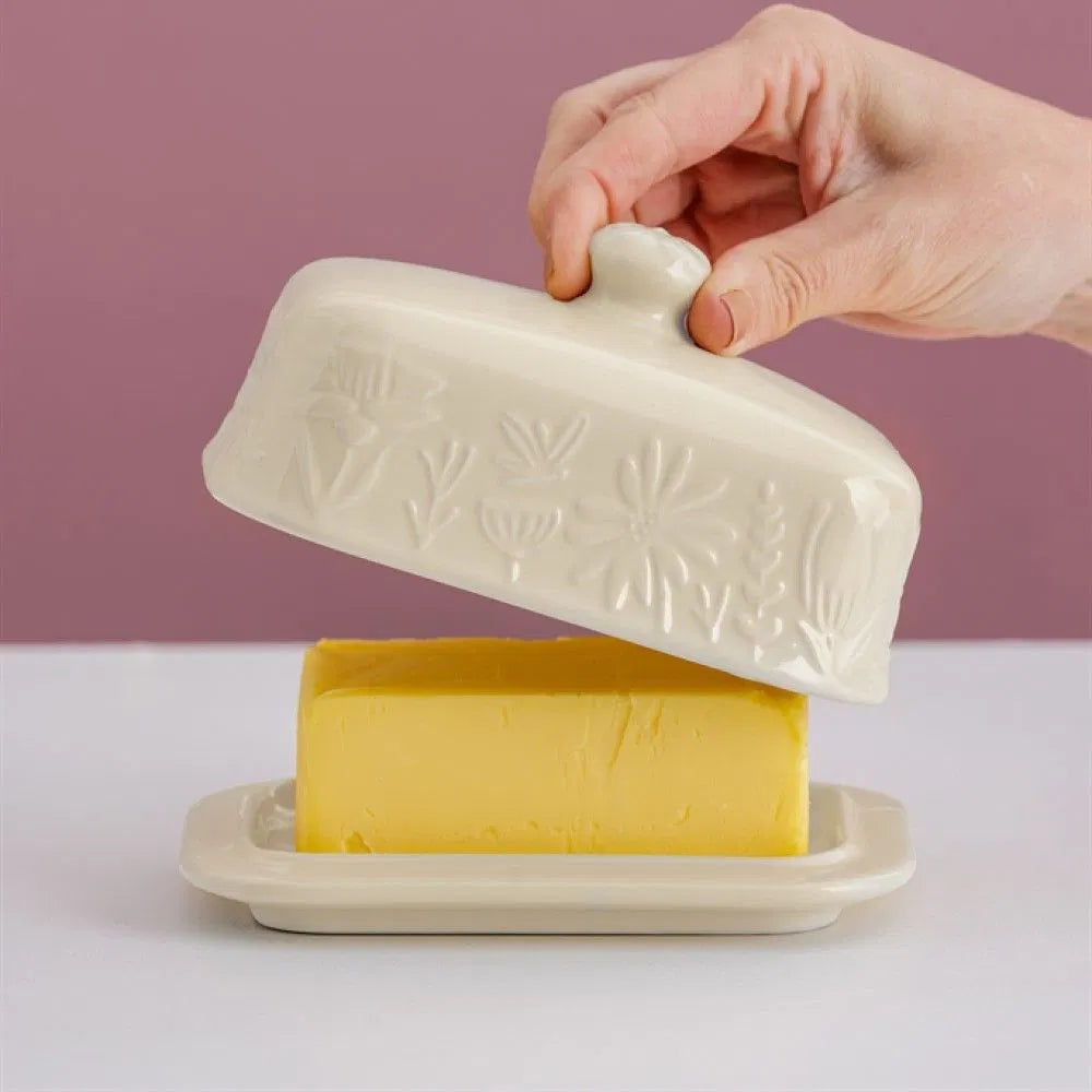 Hand opening a white ceramic butter dish with a block of butter inside on a light purple background