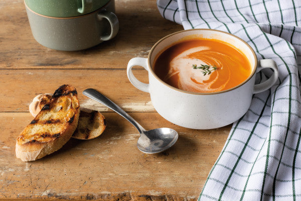 white soup mug filled with soup set on a wooden table with toasted breads, a spoon, towel, and a stack of other soup mugs in the background.