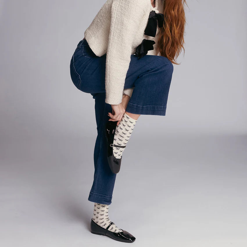 Person wearing blue jeans, a beige sweater, and black bow patterned socks on a light grey background