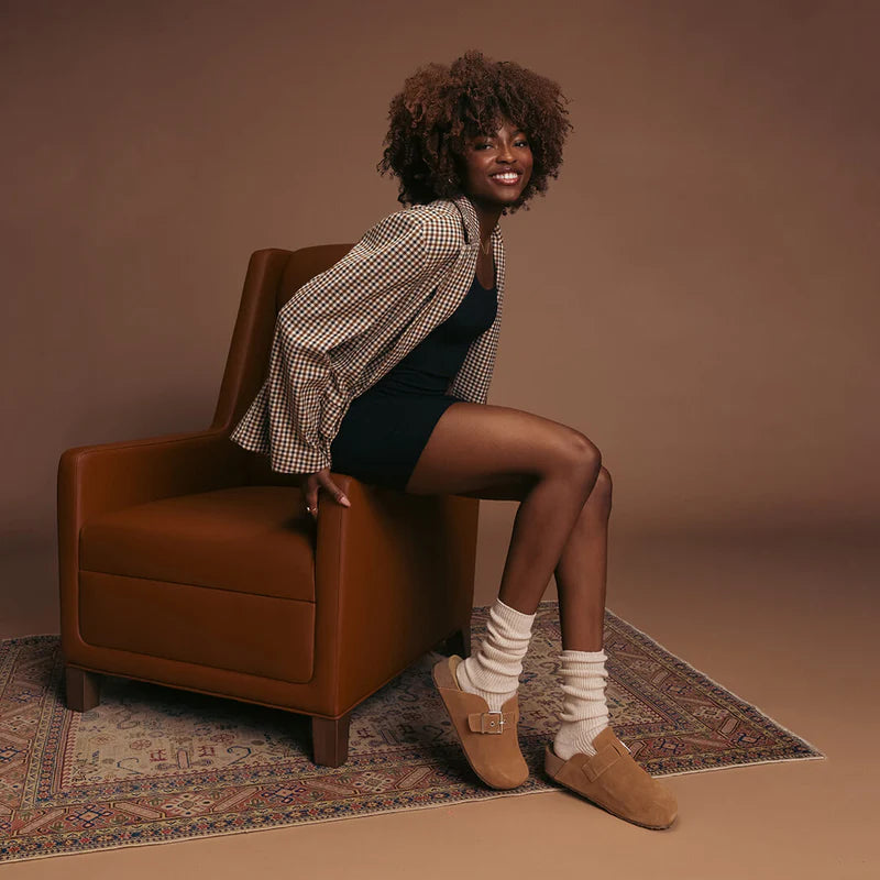 person sitting on a brown armchair wearing a checkered shirt over a black dress, ivory slouch socks, and brown shoes.