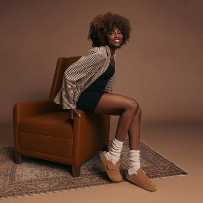 person sitting on a brown armchair wearing a checkered shirt over a black dress, ivory slouch socks, and brown shoes.
