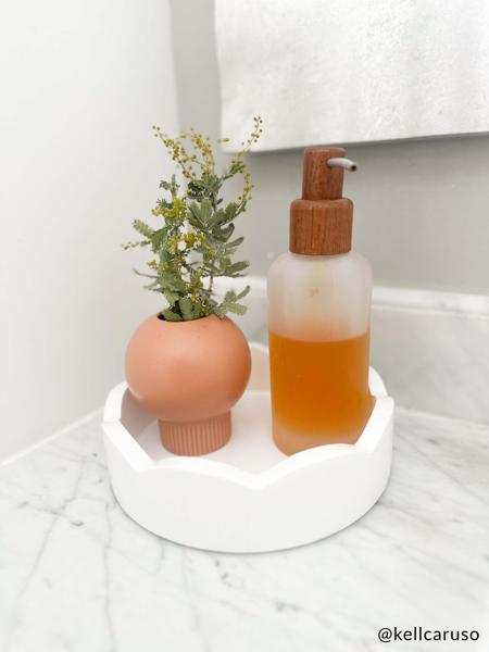Round white scalloped edge tray sat on a bathroom counter with a soap pump bottle and a small vase in it.