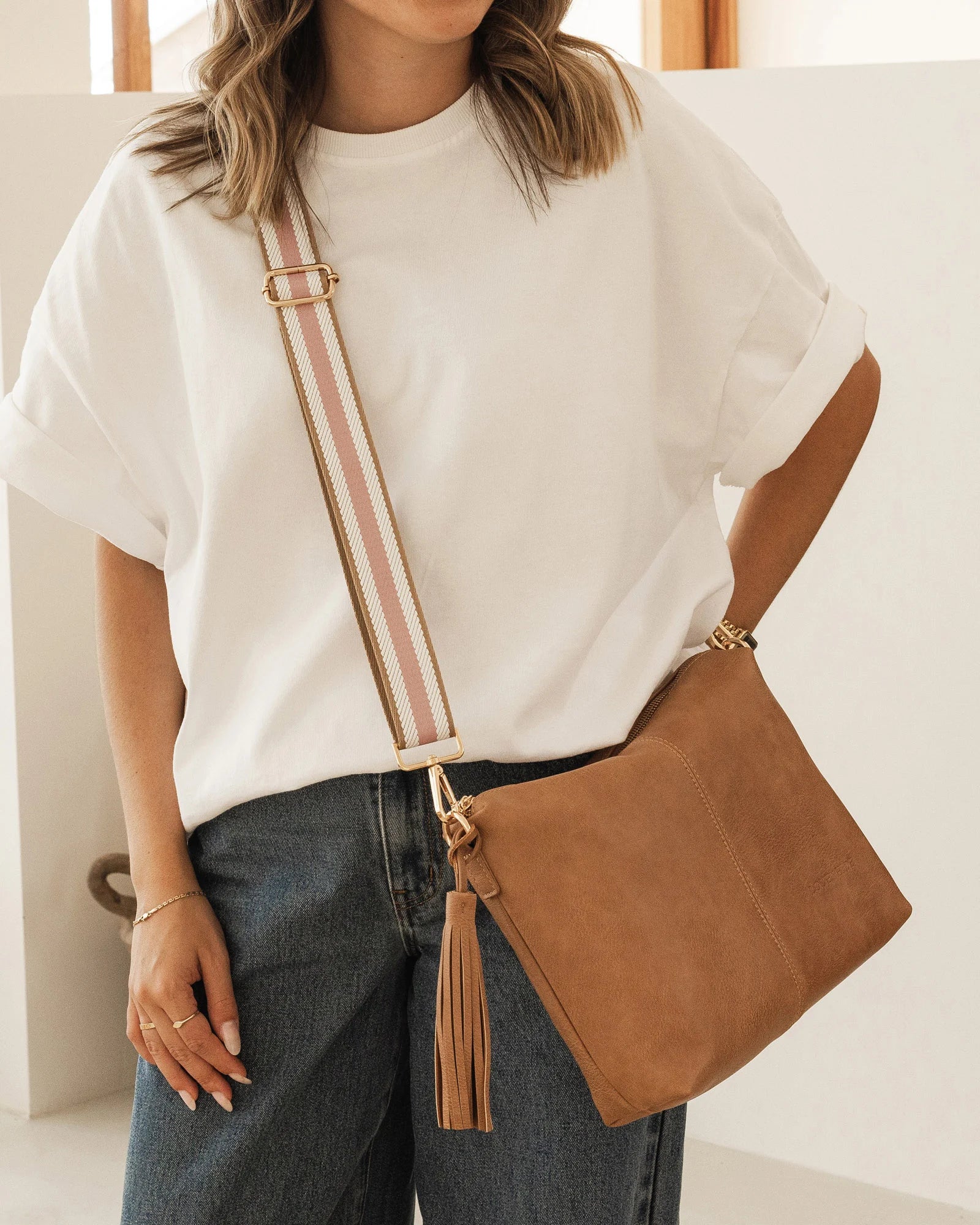 Person wearing jeans and a white T shirt Carrying camel Daisy bag with Pink stripe crossbody strap.