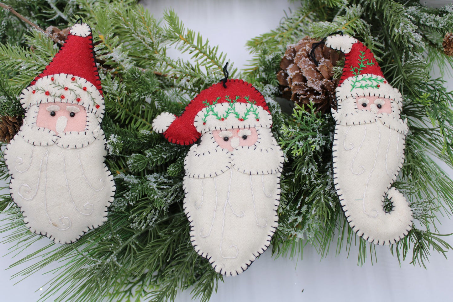 3 assortd santa face ornaments made out of felt arranged on greenery.