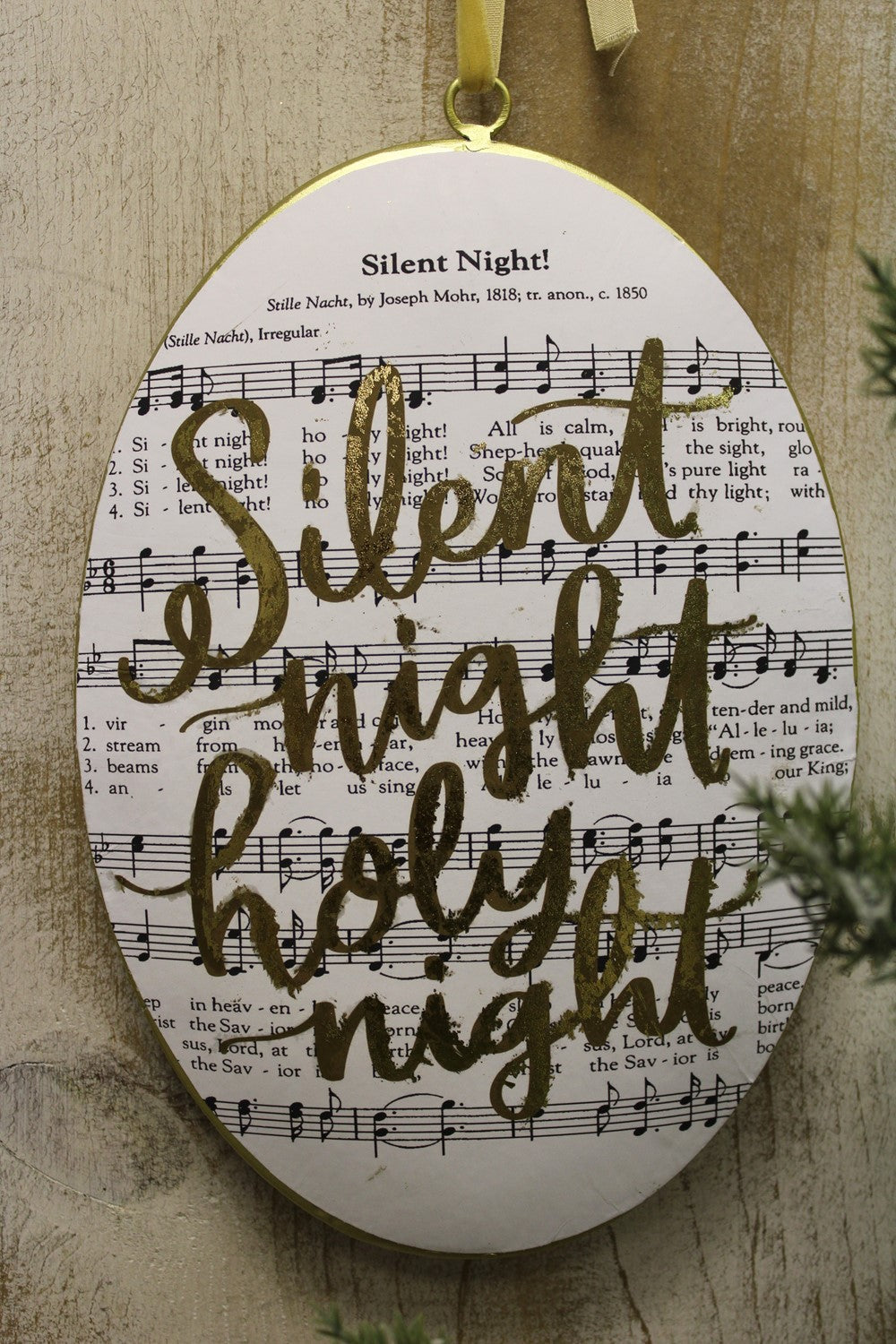 An oval Christmas ornament featuring the words 'Silent Night Holy Night' in gold script over a printed sheet music background, hanging from a gold ribbon against a wooden wall.