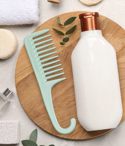Knot Today Detangling Shower Comb displayed on a wooden trivet next to bottle of conditioner, sprig of leaves, bar of soap, and towel