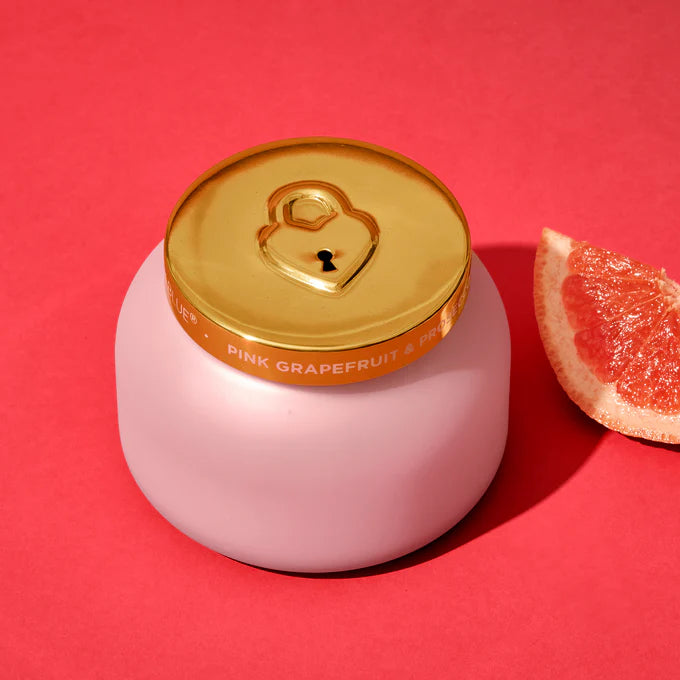 candle in a pink glass vessel with gold lid and grapefruit wedge next to it on a raspberry red background