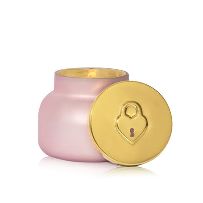 lit candle in a light pink vessel with its gold lid propped up against it