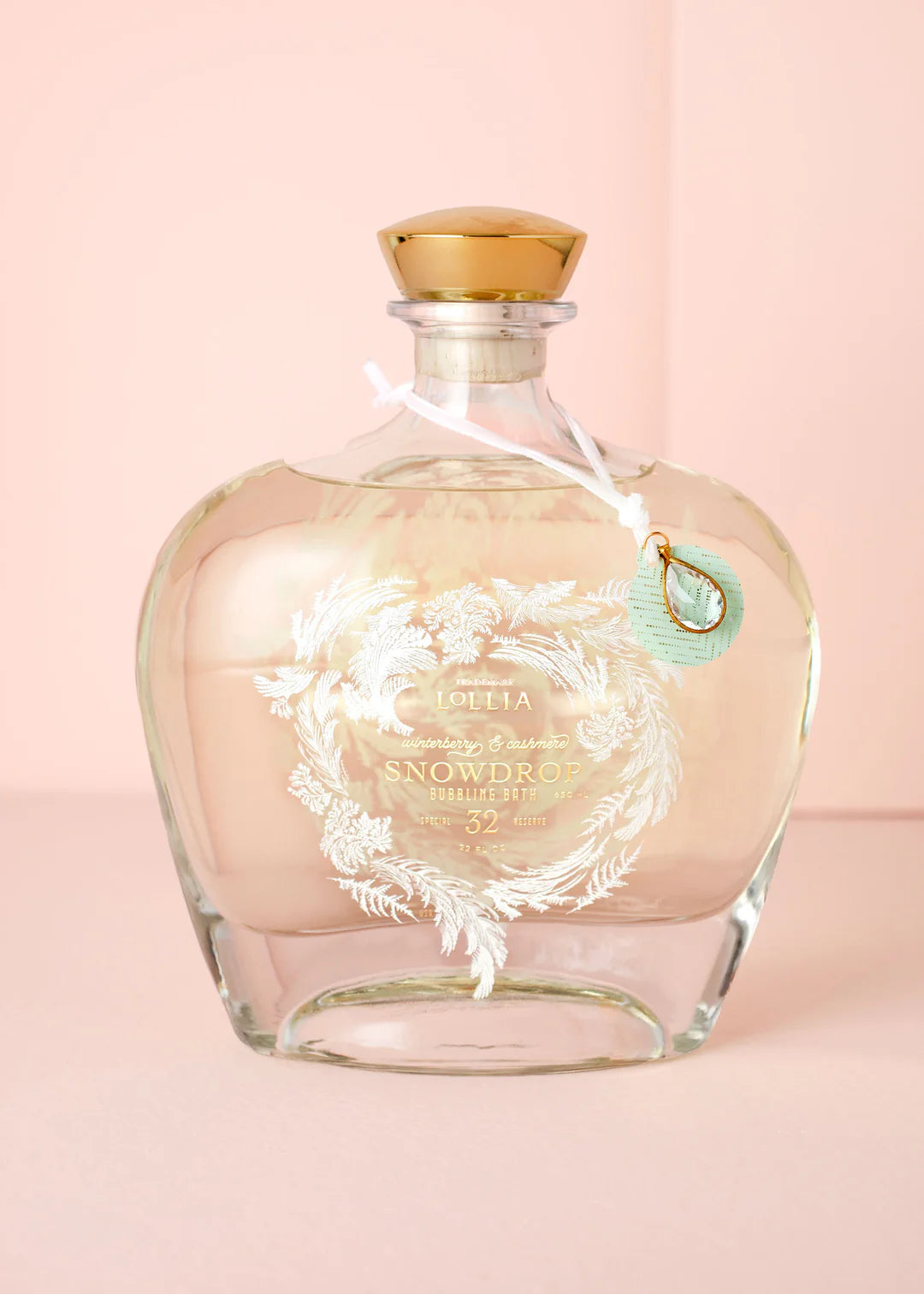 Glass bottle of snowdrop bubble bath with gold cap and decorative label on a pink background