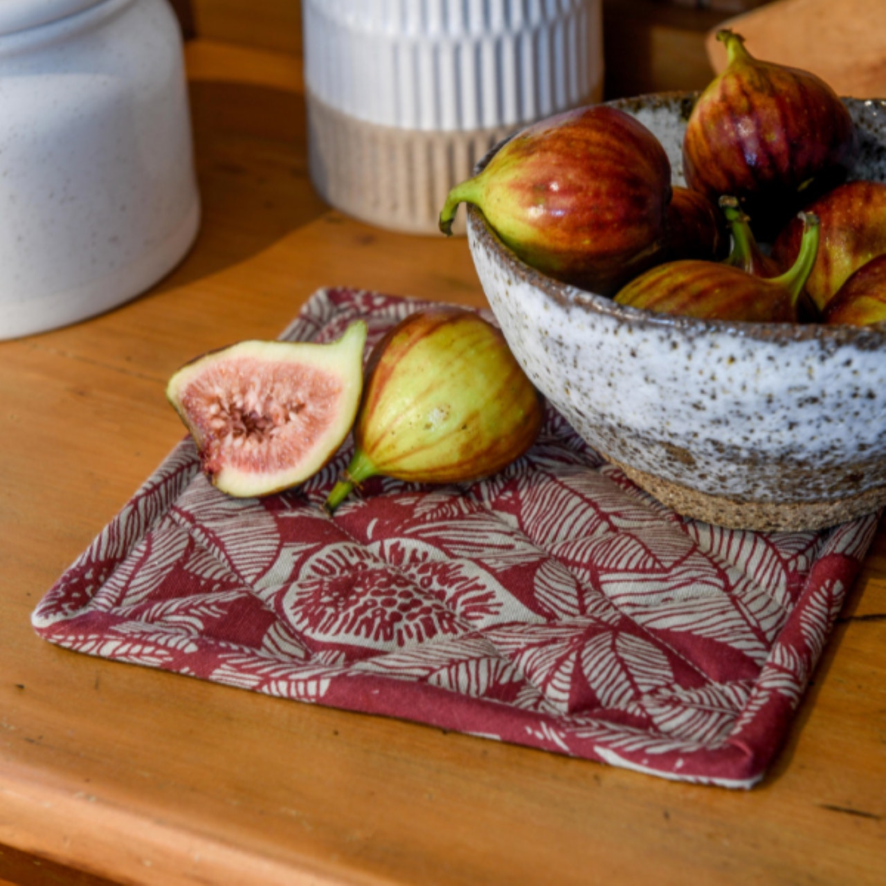 ruby and natural pot holder with fig and leaf pattern on a table with a bowl of figs set on it.