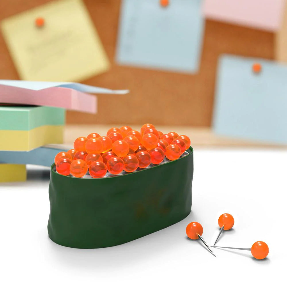 Maki Tacks Sushi Pushpins set on a desk with a stack of note papers and a bulletin board in the background.