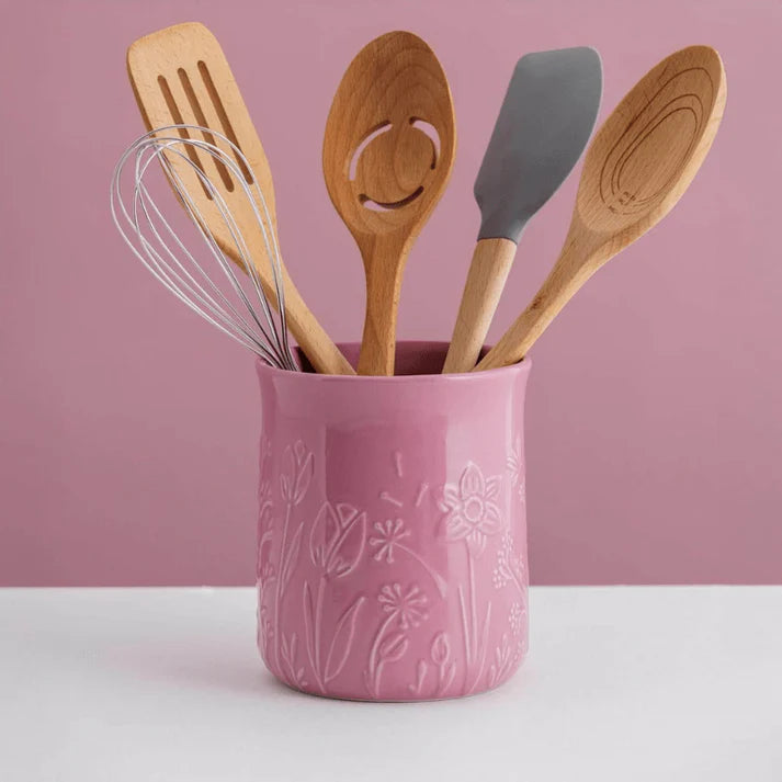 Pink ceramic utensil holder with wooden and silicone utensils in it against a pink background