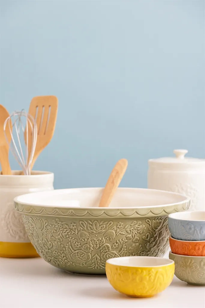home to roost pistachio green bowl with chicken and floral embossed design set on a counter with a wooden spoon in it and other bowls arranged around it