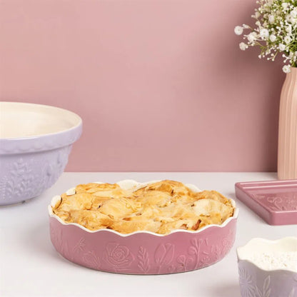 Baked pie in a pink ceramic dish on a white surface with a pink wall and decorative elements in the background.