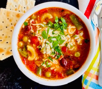Bowl of soup with pasta and vegetables, accompanied by crackers on a dark surface.