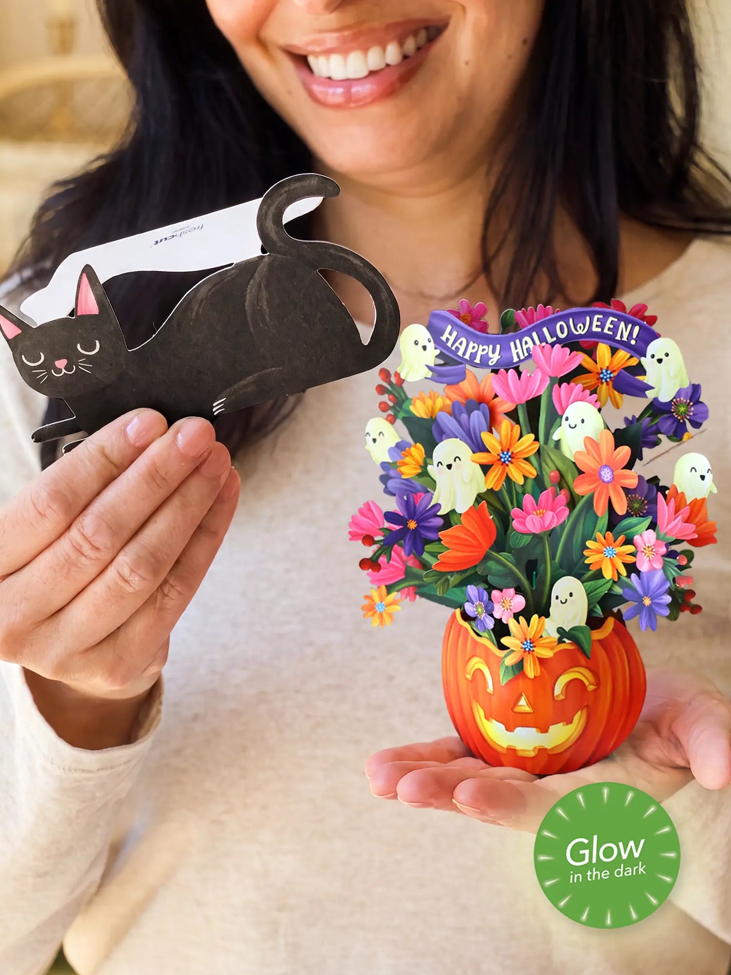 person holds a black cat-shaped card in one hand and a colorful 3D paper flower bouquet in a jack-o'-lantern pot in the other. The bouquet features small white ghosts, a purple 'Happy Halloween!' banner, and a green 'Glow in the Dark' badge overlay.