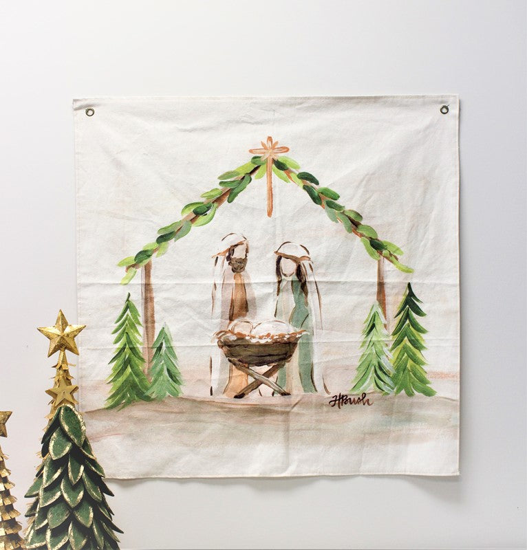 off-white fabric art with mary, joseph, and baby jesus under a creche draped with evergreens and trees on each side.