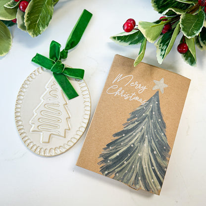 Ceramic Christmas tree ornament with green velvet bow on a white surface next to a kraft cardboard box decorated with a green christmas tree and the words "merry christmas."