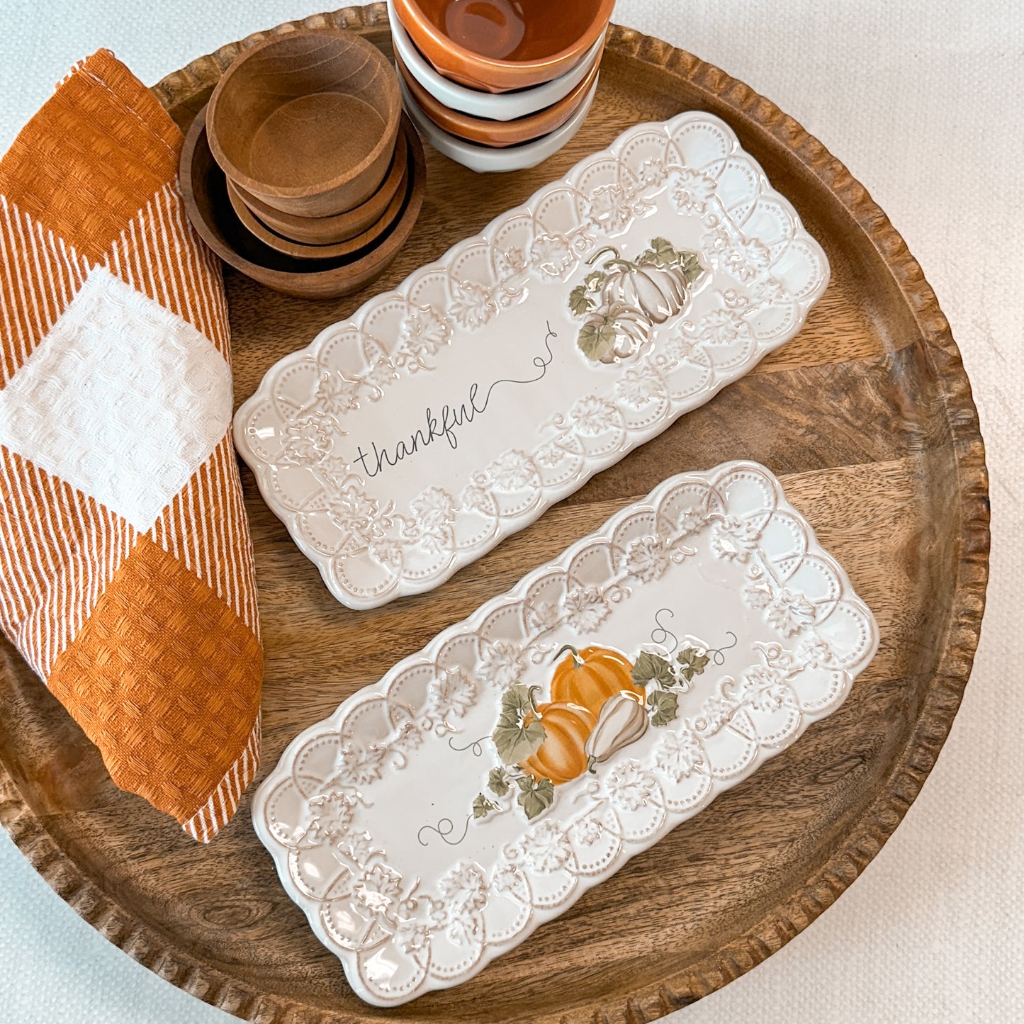 Two decorative trays with pumpkin designs one is printed with 'thankful' text next to a towel and tiny bowls.