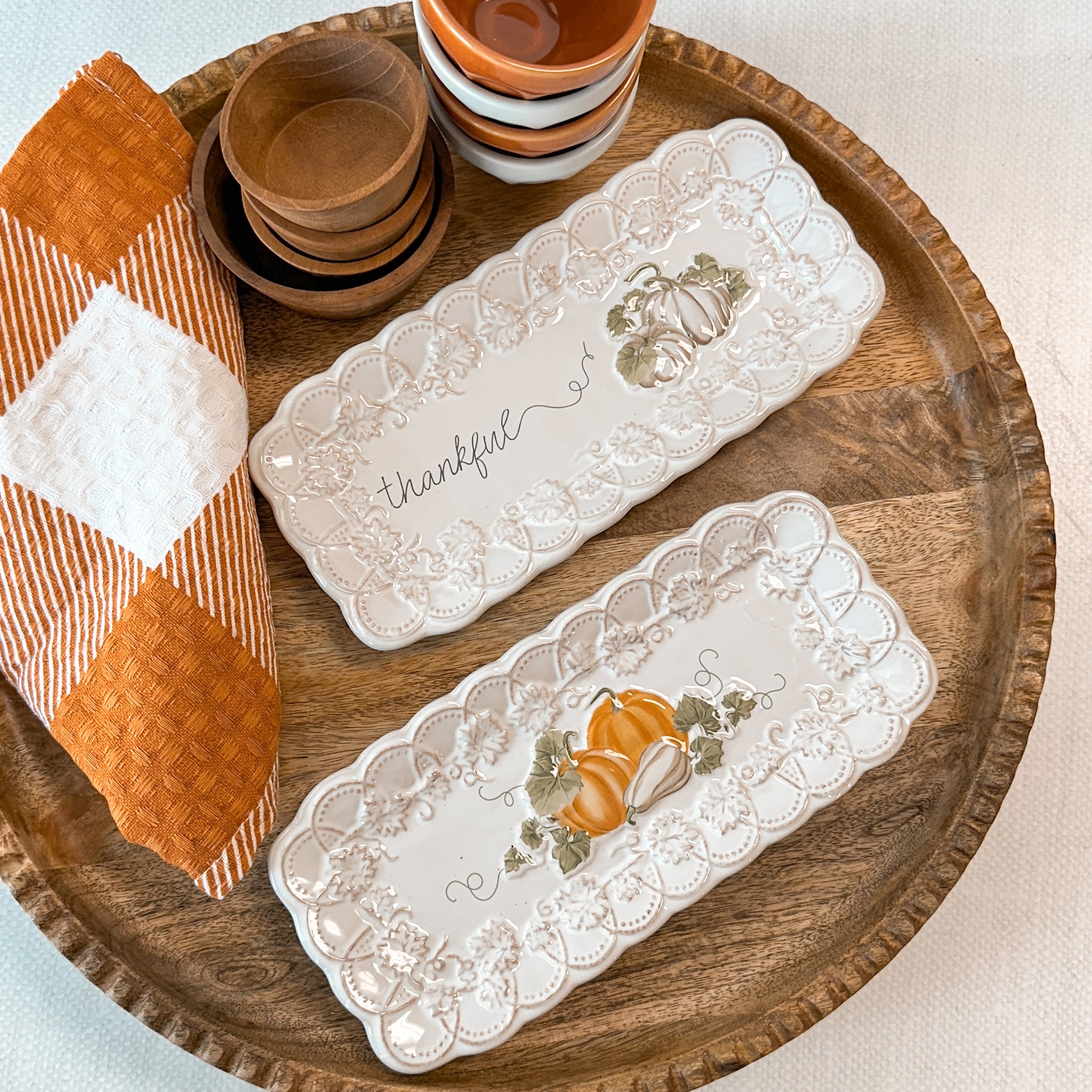 Two decorative trays with pumpkin designs one is printed with 'thankful' text next to a towel and tiny bowls.