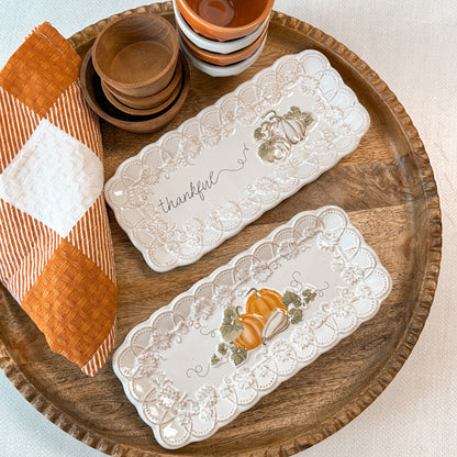 Two decorative trays with pumpkin designs one is printed with 'thankful' text next to a towel and tiny bowls.