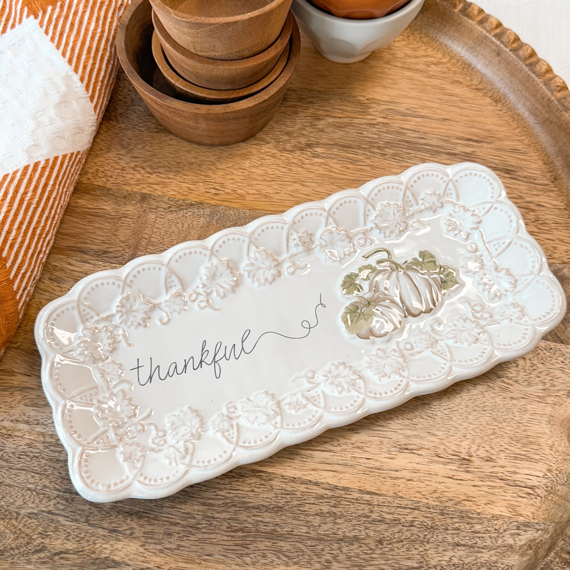 A decorative tray with pumpkin designs and printed with 'thankful' text on a wooden board with a towel and tiny bowls.