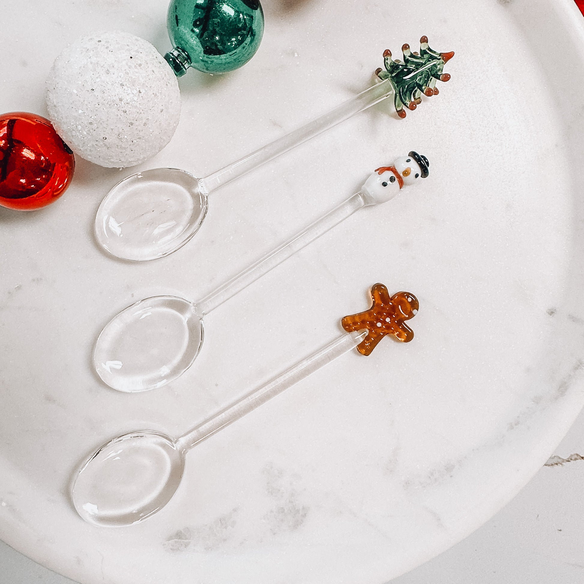 Three glass christmas spoons on a marble background, one each tree, snowman, and gingerbread.