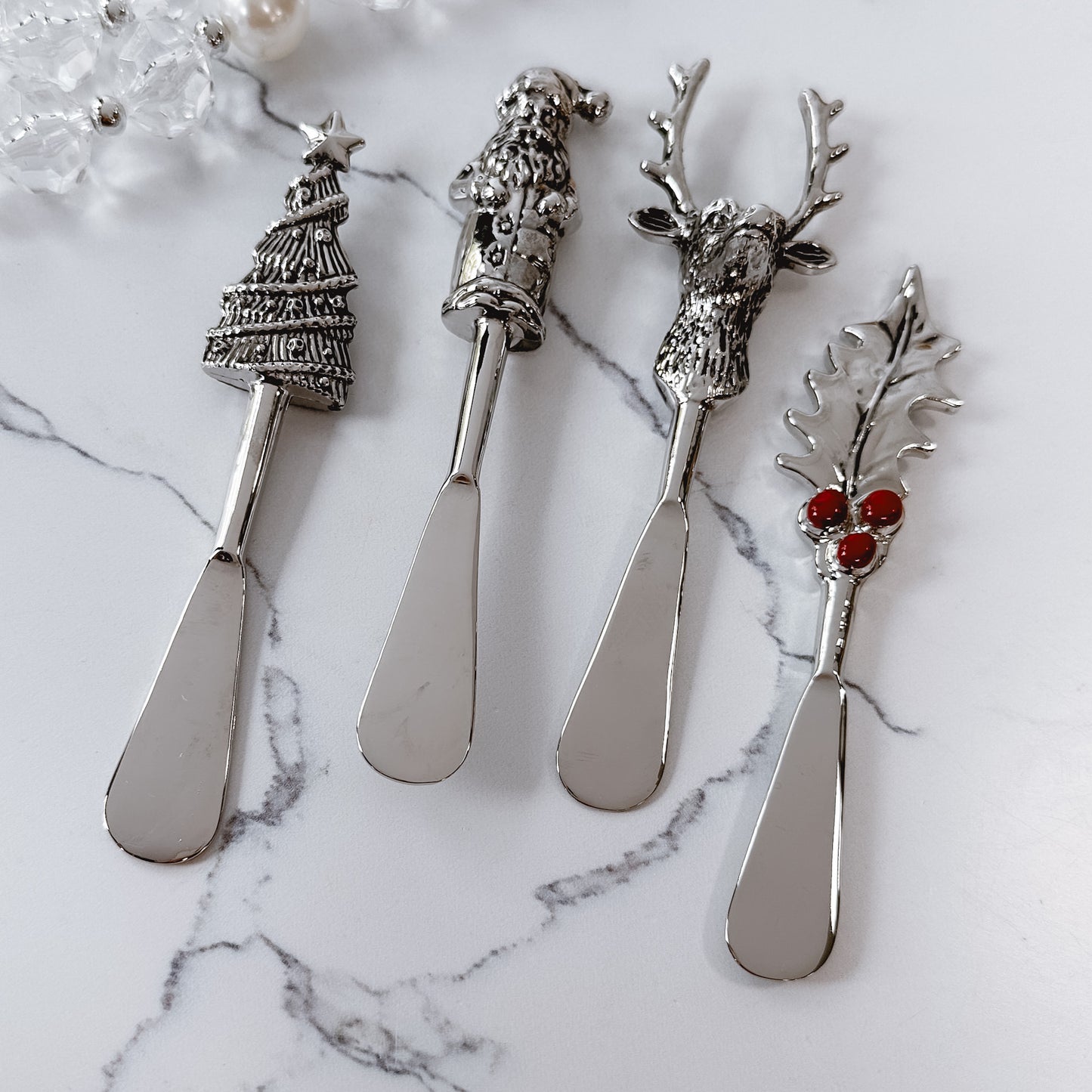 4 spreaders with assorted metal handles, one each of Santa, holly leaf, reindeer head, and a tree on a marble background.