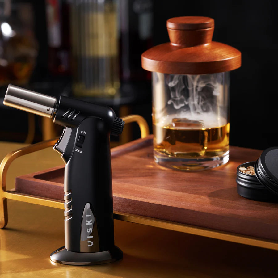 Butane torch on a wooden surface with a smoked beverage in the background