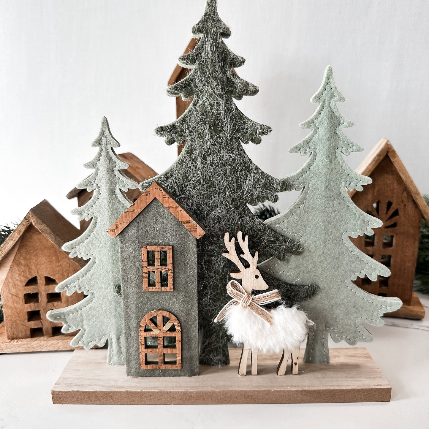Wooden base with three stand-up felt trees, a small house front, and a reindeer with floofy white body and a jute bow.