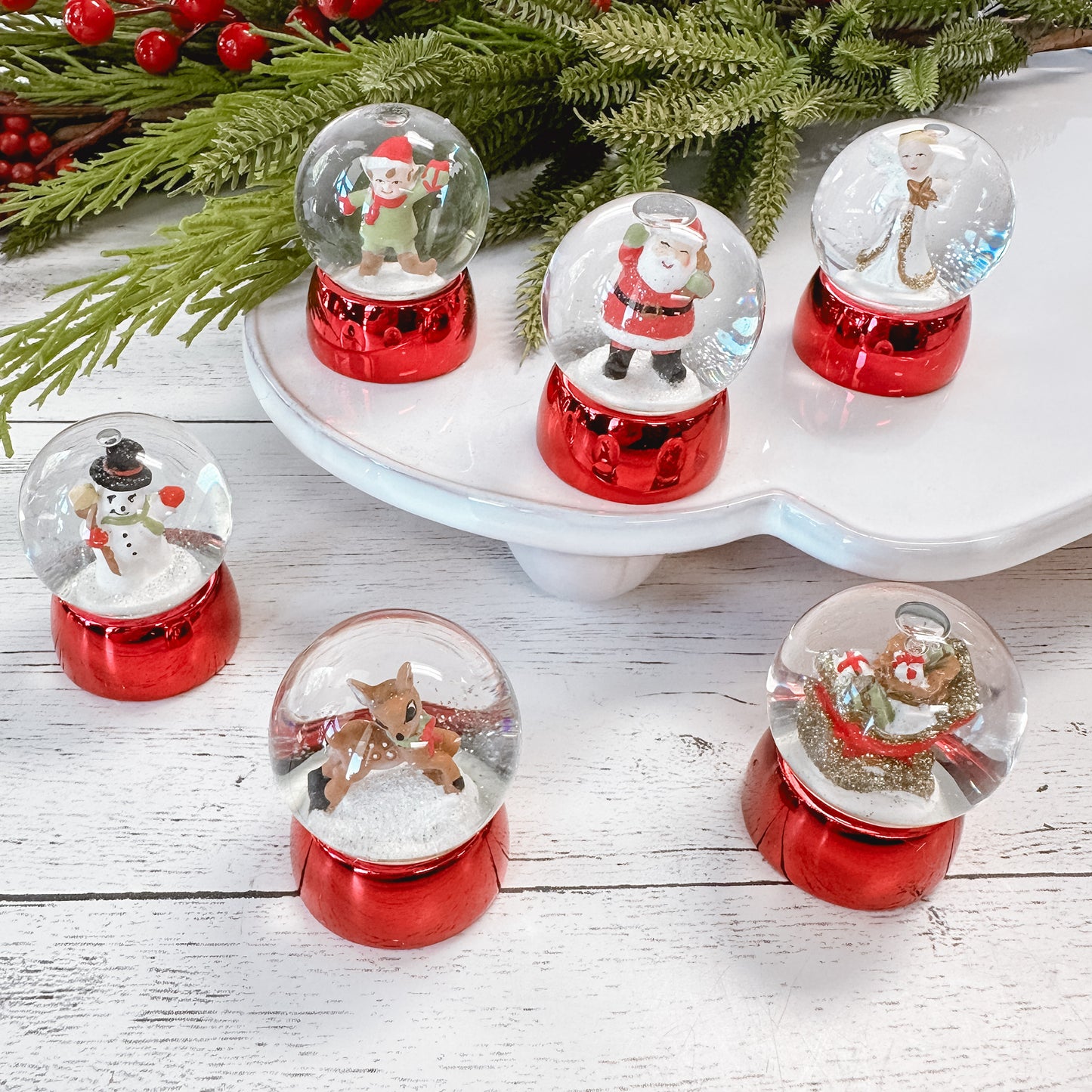 Mini water globes with red bases and a variety of Christmas characters like, santa, elf, snowman, deer, angel, and sleigh with faux foliage on a white wood tabletop.