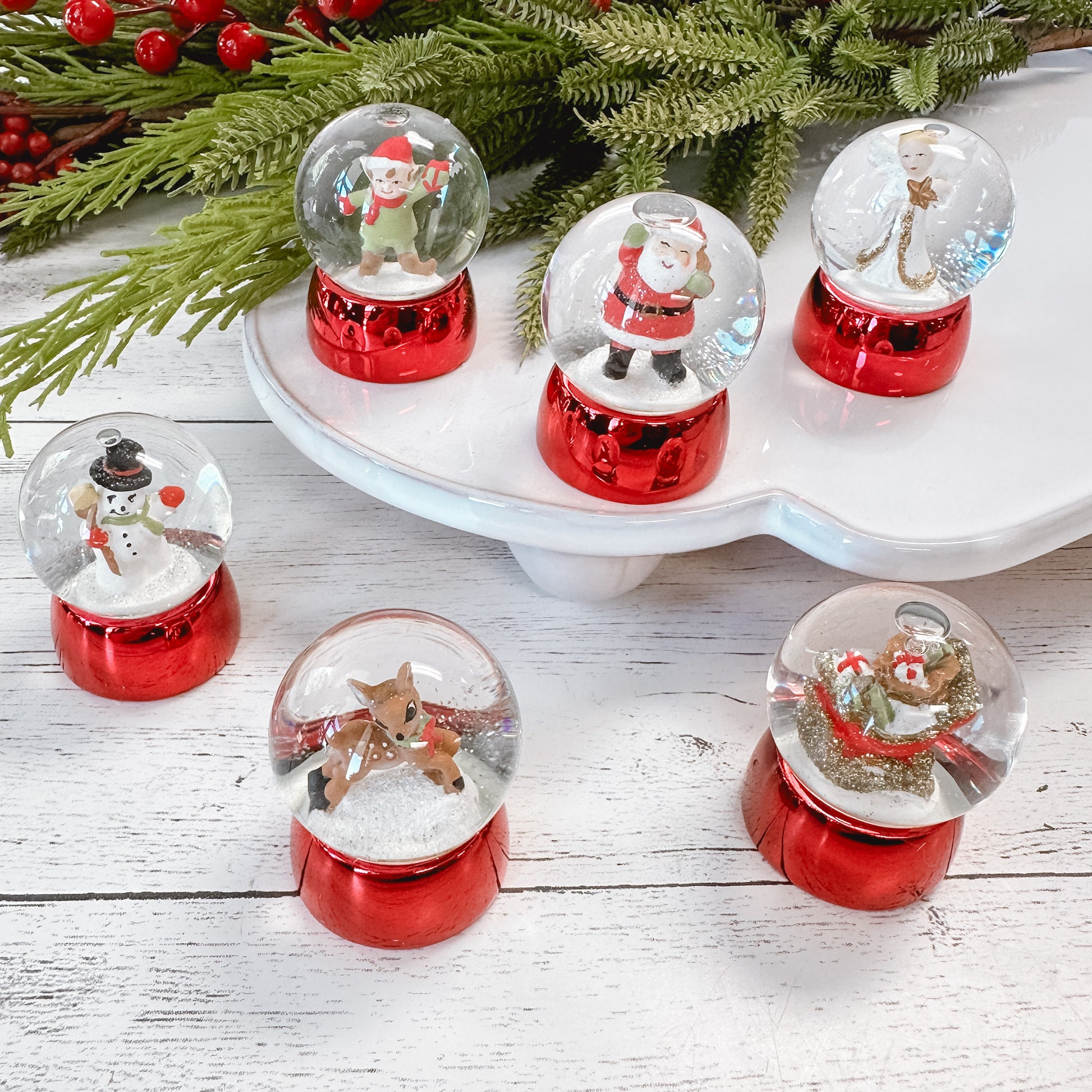 Mini water globes with red bases and a variety of Christmas characters like, santa, elf, snowman, deer, angel, and sleigh with faux foliage on a white wood tabletop.