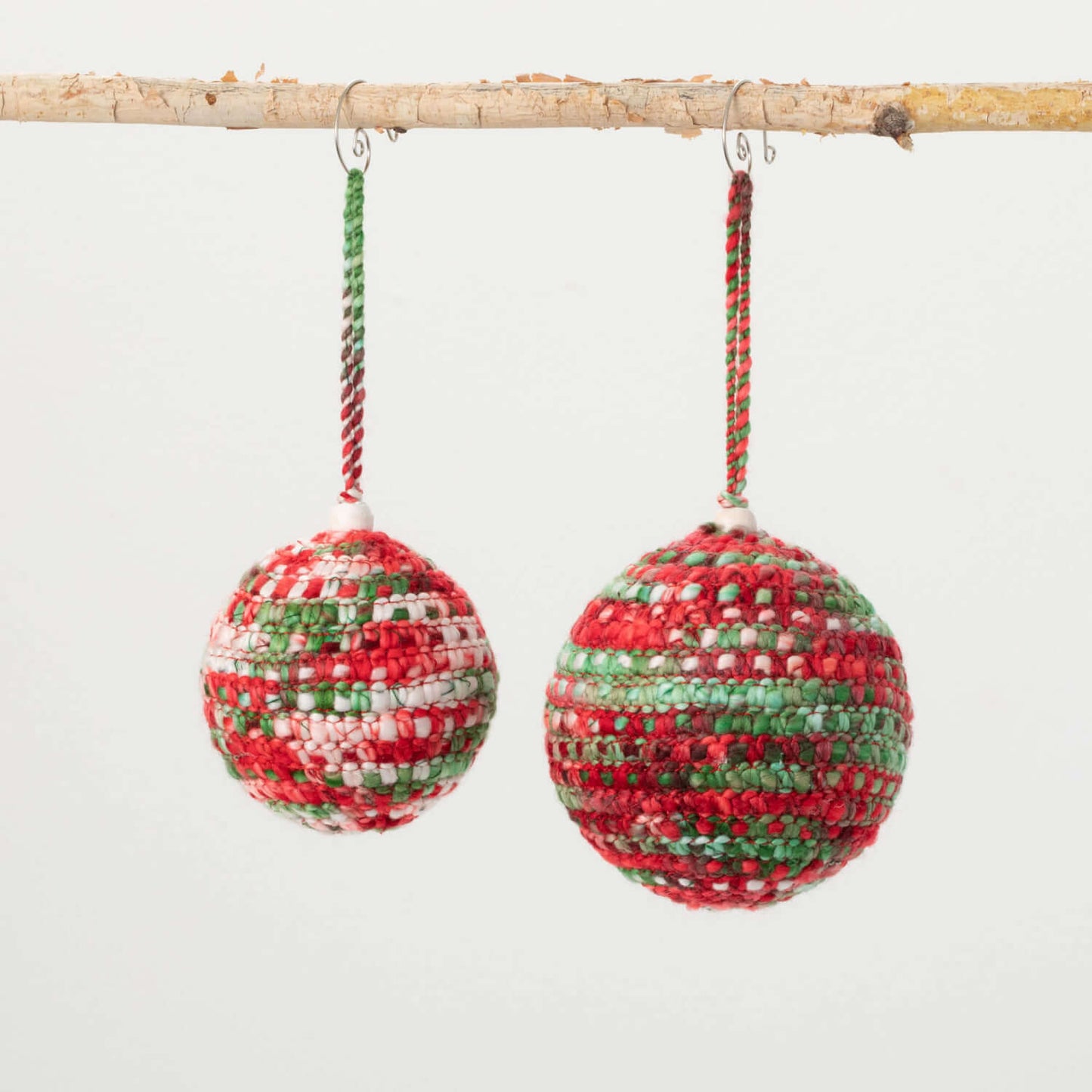 both sizes of festive knit ball ornaments hanging from a branch against a white background