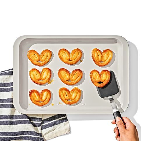 style shot of the OXO metal and ceramic quarter sheet pan with heart shaped cookies on it and a hand with spatula picking up a cookie