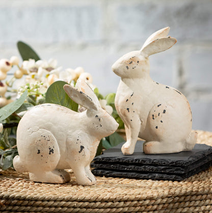 2 styles of Metal Bunny Figurines set on a basket with white floral and greenery behind them.