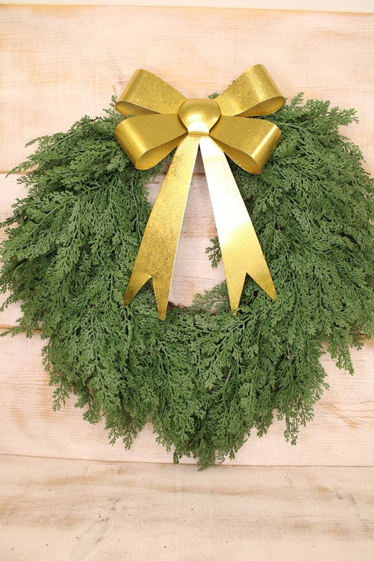 A green evergreen wreath with a large shiny gold bow on top shown on a light wood background.
