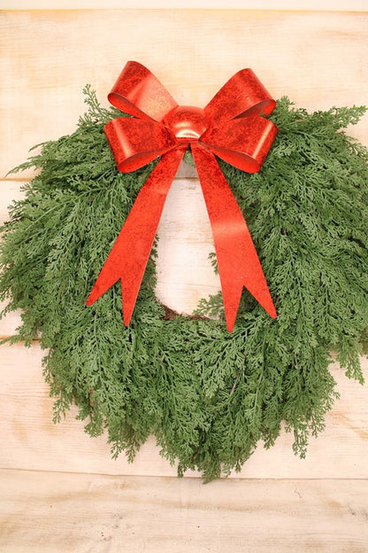 A green Christmas wreath made of dense evergreen branches, decorated with a large glittery red metal bow on the top, shown against a light wood plank background.