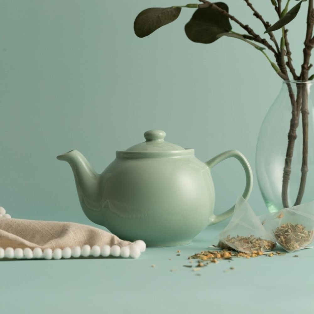 mint teapot on a light green background arranged with tea, greenery, and a cloth napkin.