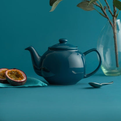 teal teapot on a blue background arranged with slices of fig, a spoon, and greenery in a vase.