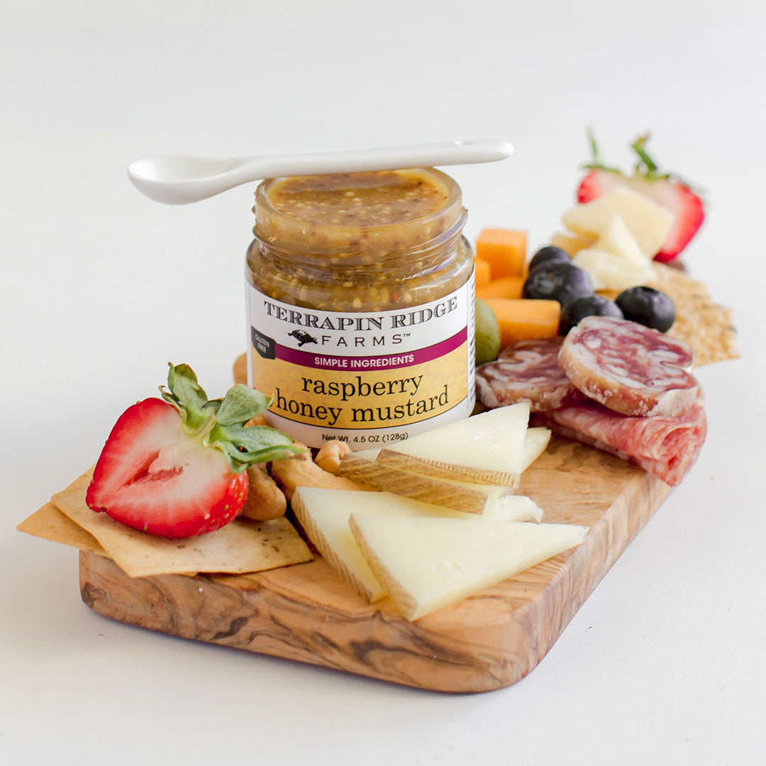 jar of Raspberry Honey Mustard on a wooden board with meats, cheeses, and fruit.