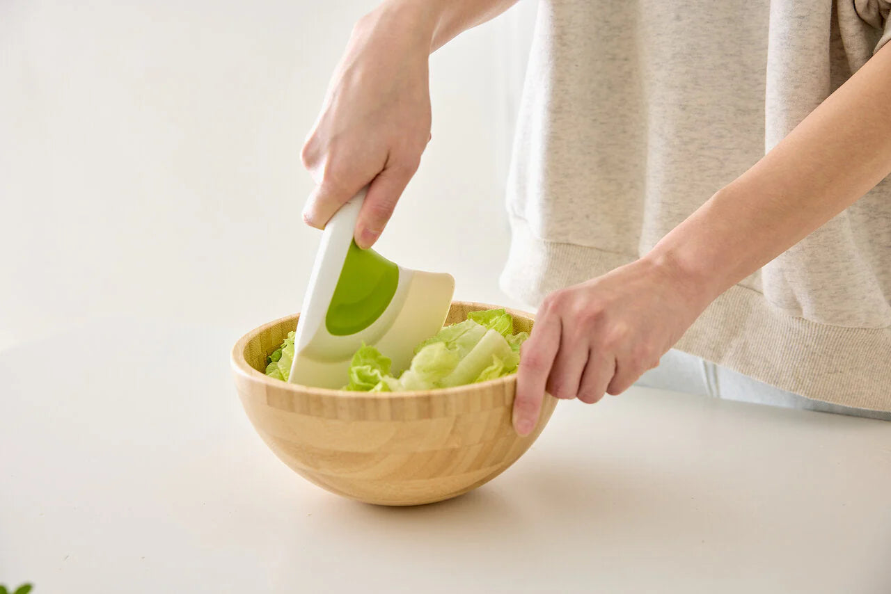 Person using a green kitchen tool to chop vegetables in a wooden bowl on a light surface.