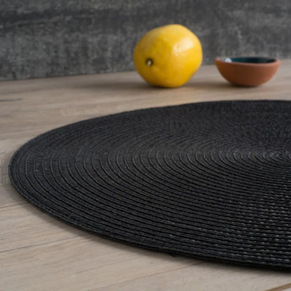 Black round placemat on a wooden table with a lemon and a bowl in the background.