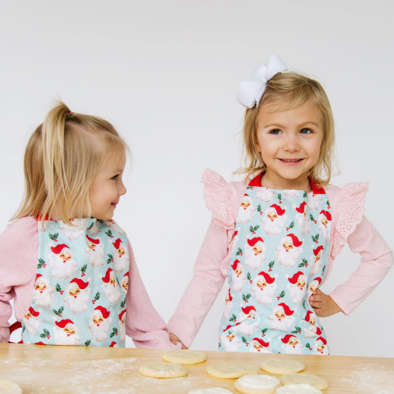 2 children wearing santa aprons standing at a counter filled with cookies.