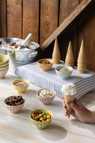 hand holding ice cream cone with assorted ice cream bowls filled with toppings in the background.