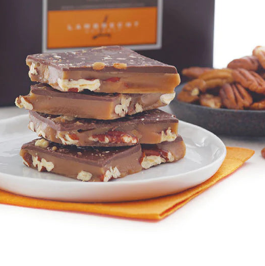 Stack of chocolate-covered toffee pieces on a white plate with a bowl of pecans in the background, featuring the Lamberts brand.