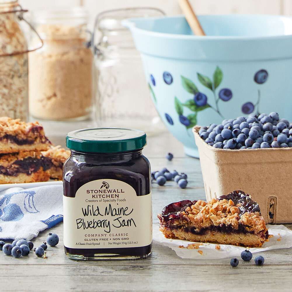 jar of Wild Maine Blueberry Jam set on a table with a pastry, a basket of blueberries, and and mixing bowl.