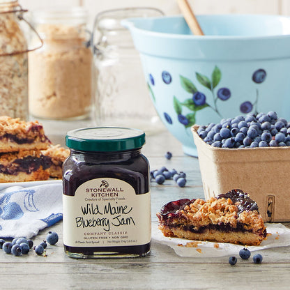 jar of Wild Maine Blueberry Jam set on a table with a pastry, a basket of blueberries, and and mixing bowl.